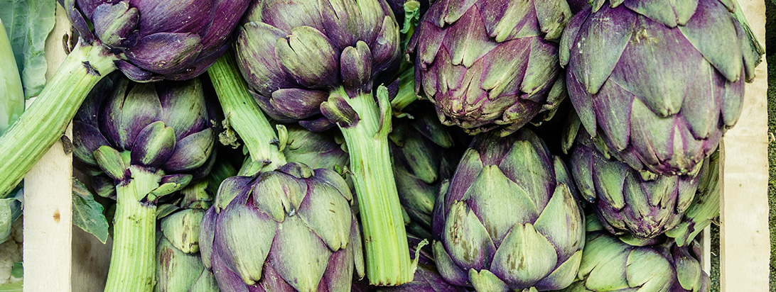 Numerous fresh purple and green artichokes with long green stems are piled together in a crate.
